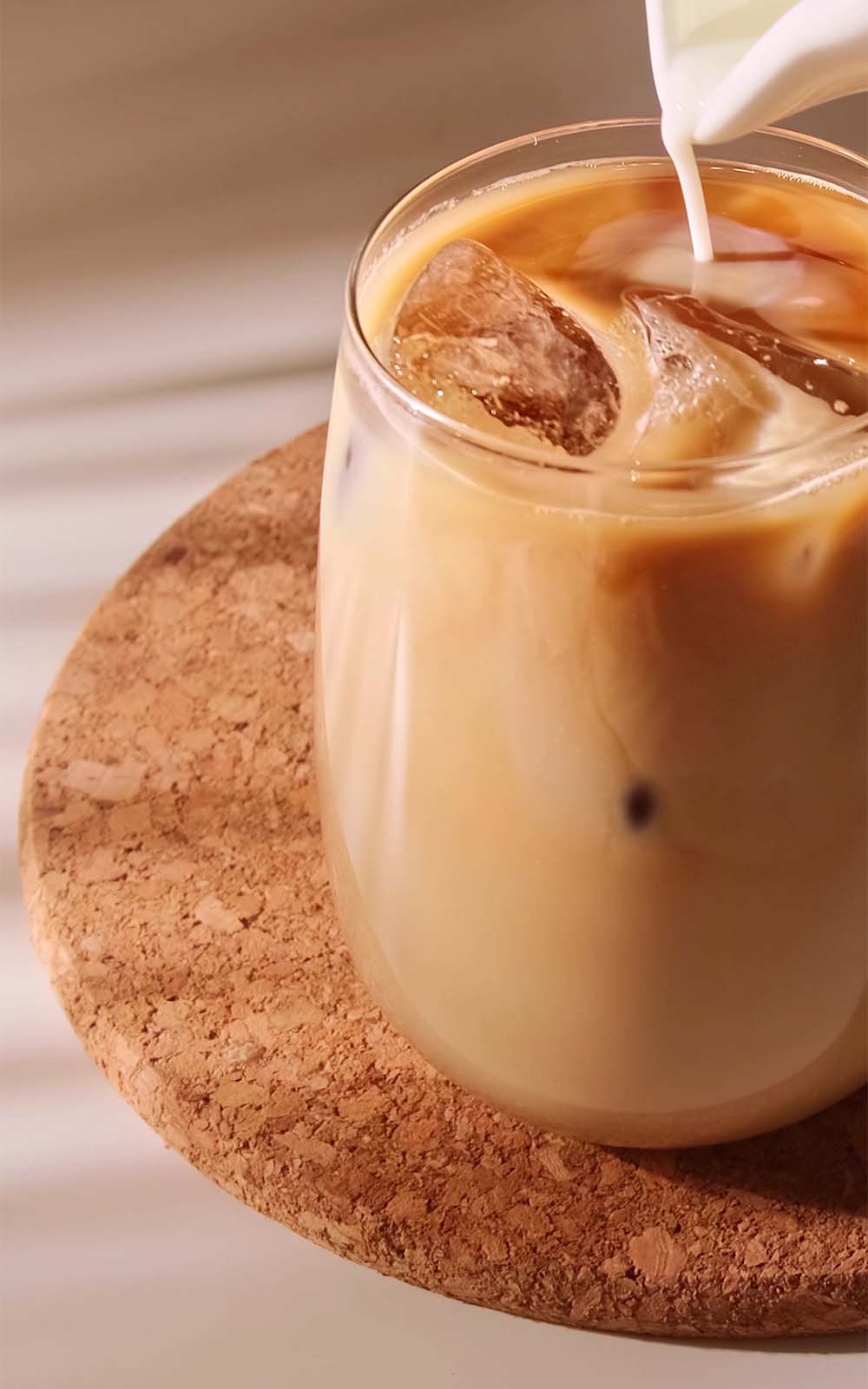 Glas mit Eiskaffee und Eiswürfeln, auf Korkuntersetzer stehend, während von oben Milch eingegossen wird. Glas mit Eiskaffee und Eiswürfeln, auf Korkuntersetzer stehend, während von oben Milch eingegossen wird.