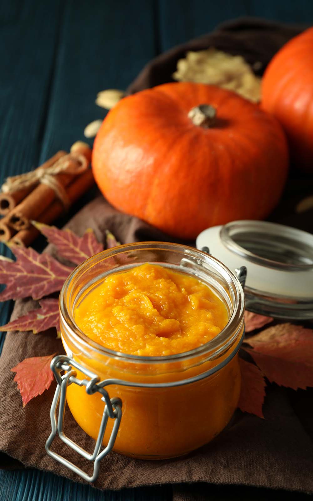 Ein Glas mit cremigem orangefarbenem Kürbisaufstrich, dekoriert mit herbstlichen Blättern und Zimtstangen. Im Hintergrund liegen ganze Kürbisse, die eine herbstliche Stimmung erzeugen. Ein Glas mit cremigem orangefarbenem Kürbisaufstrich, dekoriert mit herbstlichen Blättern und Zimtstangen. Im Hintergrund liegen ganze Kürbisse, die eine herbstliche Stimmung erzeugen.