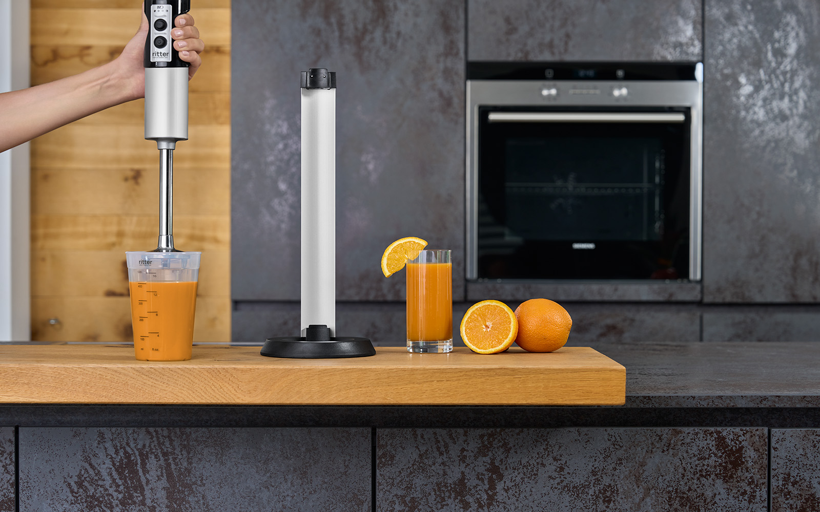 Ein Bild eines silbernen Stabmixers, der Orangen im Messbecher mixt und in einer Küche, neben seiner Ladestation, steht.