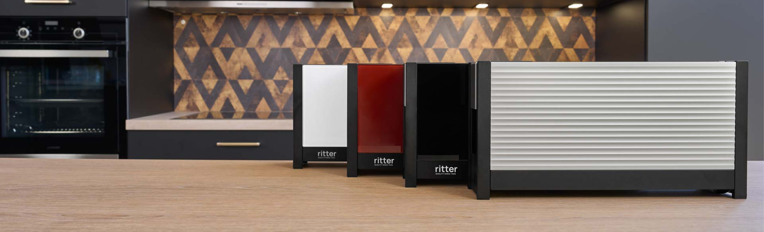 Ein Bild von einem Aluminium-, einem weißen, einem roten und einem schwarzen Toaster, die aufgereiht , hintereinander, in einer Küche stehen.