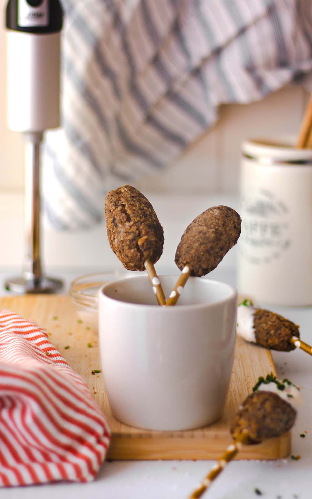 Champignon Party-Frikadellen auf kleinen Sticks serviert, in einer weißen Tasse angerichtet. Im Hintergrund sind Küchenutensilien und ein ritter Stabmixer zu sehen