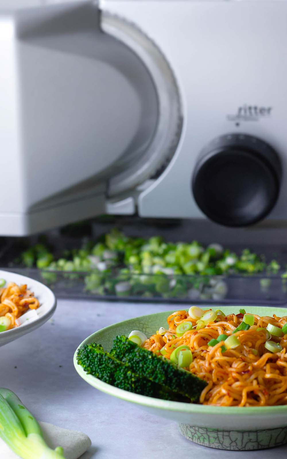Grüne Schale mit Spicy Garlic Noodles, garniert mit Frühlingszwiebeln und Brokkoliröschen. Im Hintergrund eine Küchenmaschine und frisch geschnittene Lauchzwiebeln