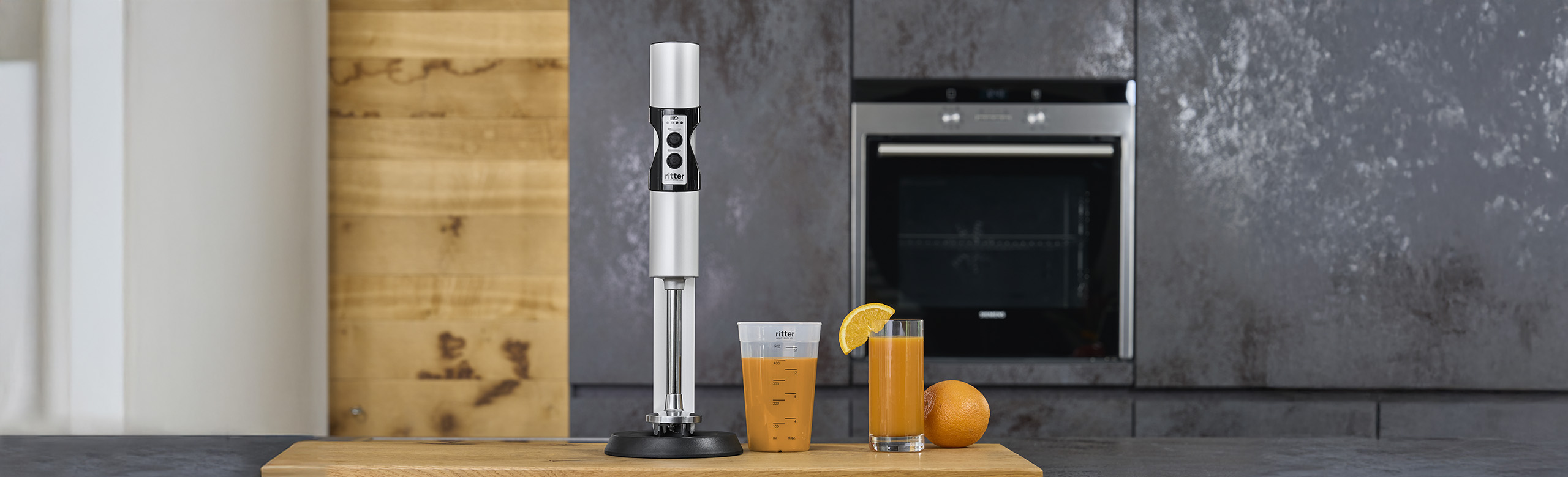 A picture of a silver hand blender, a filled measuring jug and a glass filled with juice in a kitchen.