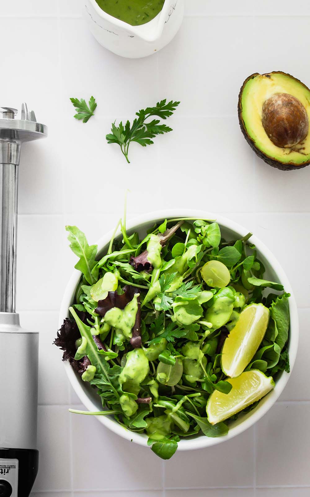Frischer Sommersalat mit grünem Blattsalat, Trauben und Avocado-Dressing in einer weißen Schüssel, daneben Limettenspalten, ein Pürierstab, frische Kräuter und eine halbe Avocado. Frischer Sommersalat mit grünem Blattsalat, Trauben und Avocado-Dressing in einer weißen Schüssel, daneben Limettenspalten, ein Pürierstab, frische Kräuter und eine halbe Avocado.