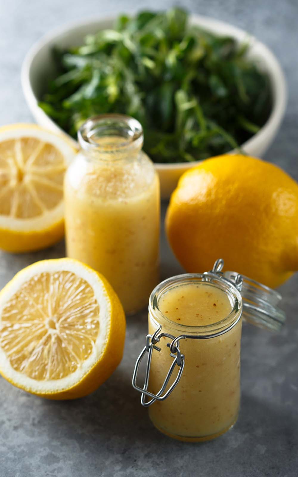 Ein Glas mit cremigem gelbem Sommer-Dressing, daneben frische Zitronenhälften und eine weitere Flasche Dressing. Im Hintergrund steht eine Schüssel mit frischem grünen Salat. Ein Glas mit cremigem gelbem Sommer-Dressing, daneben frische Zitronenhälften und eine weitere Flasche Dressing. Im Hintergrund steht eine Schüssel mit frischem grünen Salat.