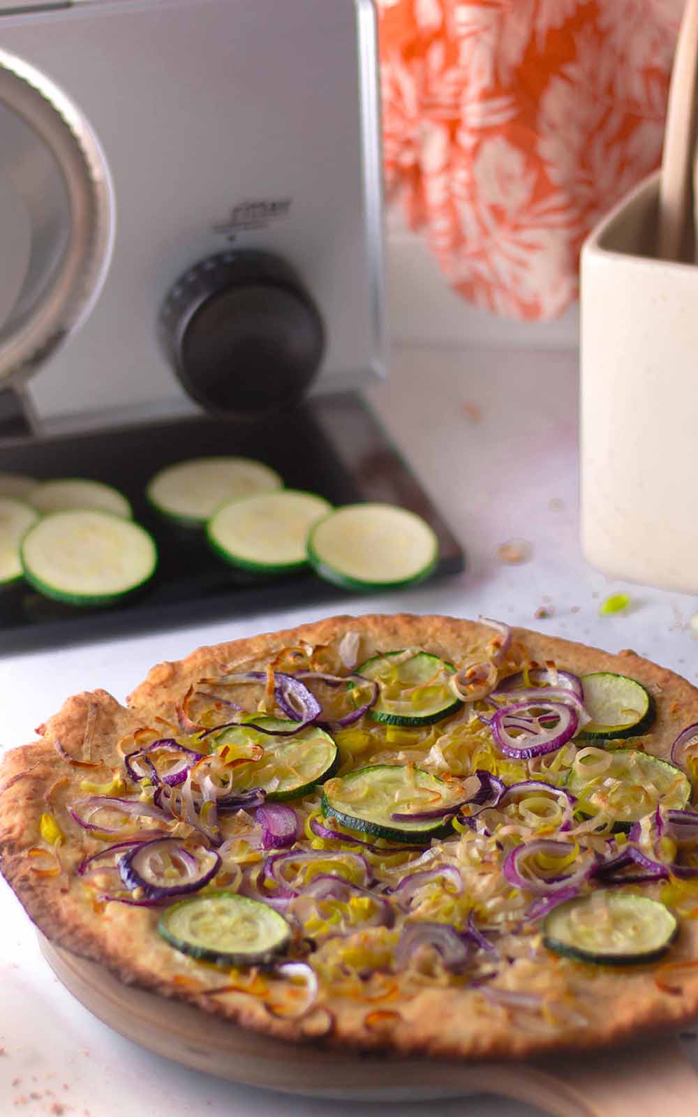 Frisch gebackene Veggie-Knoblauch-Pizza mit Zucchini- und roten Zwiebelscheiben, serviert auf einem Holzbrett. Im Hintergrund eine Küchenmaschine und einige geschnittene Zucchinischeiben