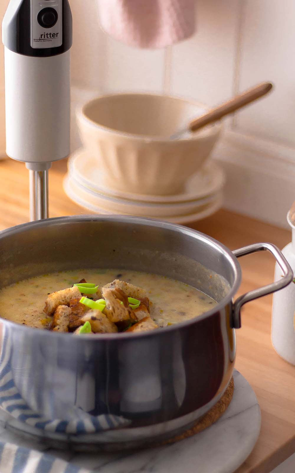 Kartoffel-Lauch-Suppe in einem Kochtopf, garniert mit knusprigen Croutons und grünen Lauchstückchen, daneben ein Stabmixer in der Küche. Kartoffel-Lauch-Suppe in einem Kochtopf, garniert mit knusprigen Croutons und grünen Lauchstückchen, daneben ein Stabmixer in der Küche.