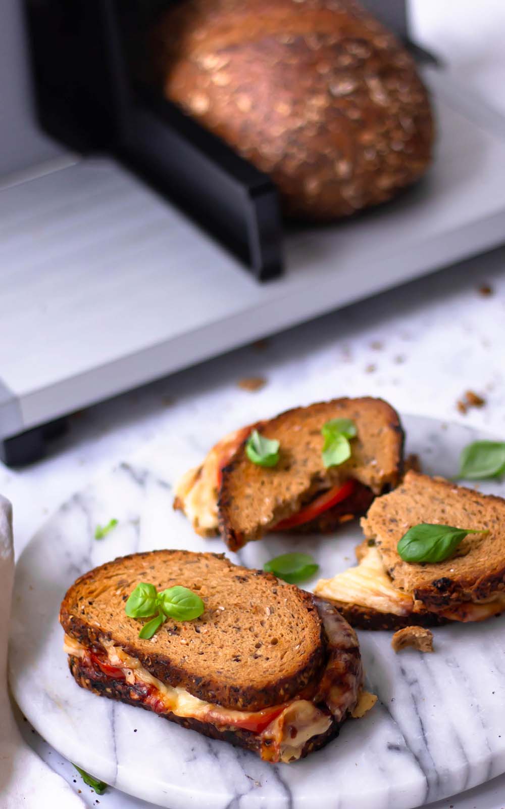 Kross gebratenes Vollkorn-Sandwich mit geschmolzenem Käse und frischen Tomatenscheiben, garniert mit Basilikumblättern, serviert auf einer runden Marmorplatte. Kross gebratenes Vollkorn-Sandwich mit geschmolzenem Käse und frischen Tomatenscheiben, garniert mit Basilikumblättern, serviert auf einer runden Marmorplatte.