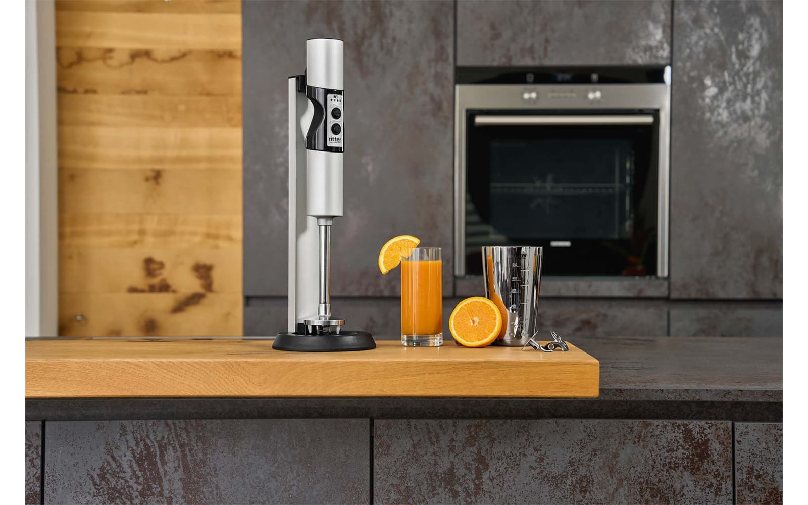 Ein Bild eines silbernen Stabmixers der neben einem Glas Orangensaft, in einer Küche steht.
