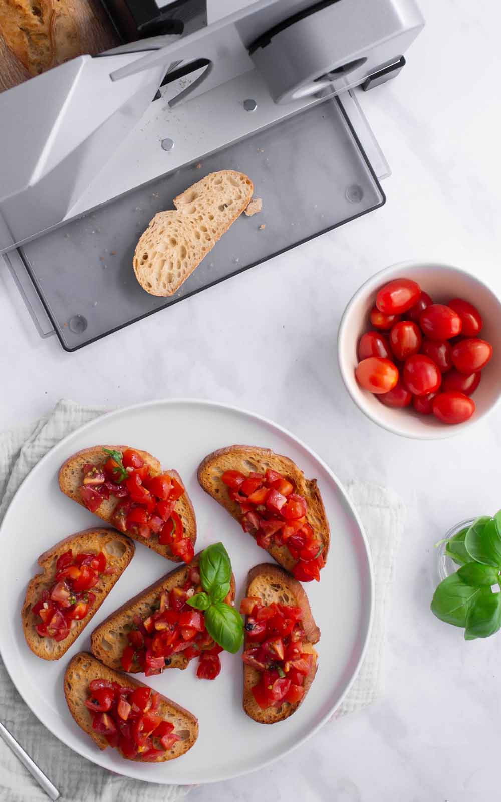 Frische Bruschetta mit Tomatenwürfeln und Basilikum auf gerösteten Brotscheiben, angerichtet auf einem weißen Teller vor einem ritter Allesschneider mit frisch geschnittenem Baguette Frische Bruschetta mit Tomatenwürfeln und Basilikum auf gerösteten Brotscheiben, angerichtet auf einem weißen Teller vor einem ritter Allesschneider mit frisch geschnittenem Baguette