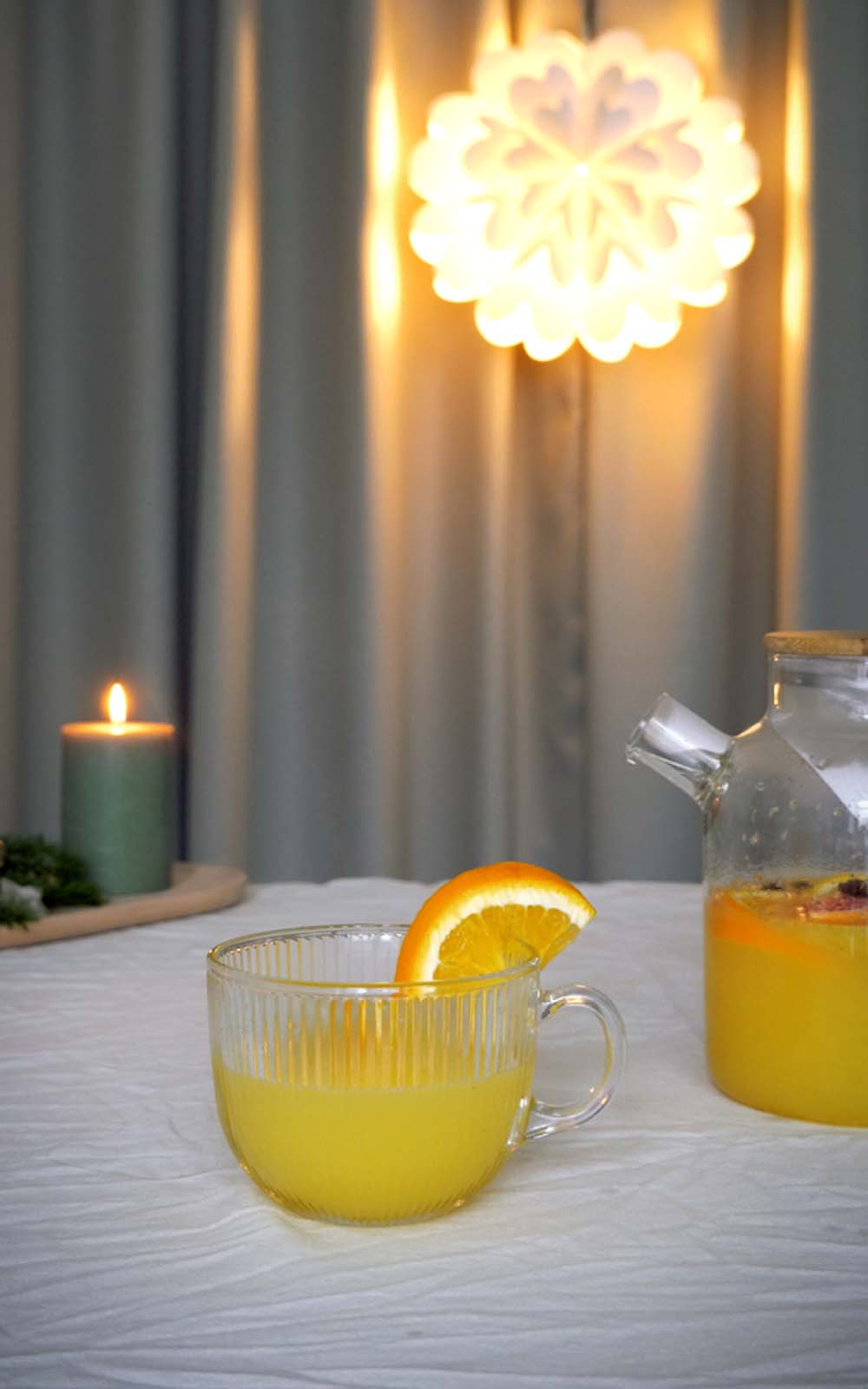 Glas mit winterlichem Zitrus-Punsch, garniert mit einer Orangenscheibe, daneben eine Glaskaraffe mit dem Punsch; im Hintergrund brennende Kerze und warme Lichter. Glas mit winterlichem Zitrus-Punsch, garniert mit einer Orangenscheibe, daneben eine Glaskaraffe mit dem Punsch; im Hintergrund brennende Kerze und warme Lichter.
