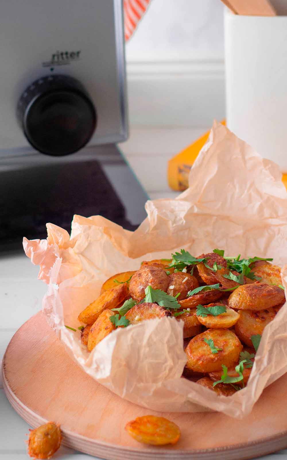 Knusprige Country Potatoes in Backpapier serviert, garniert mit frischer Petersilie. Im Hintergrund ist ein ritter Allesschneider zu sehen Knusprige Country Potatoes in Backpapier serviert, garniert mit frischer Petersilie. Im Hintergrund ist ein ritter Allesschneider zu sehen