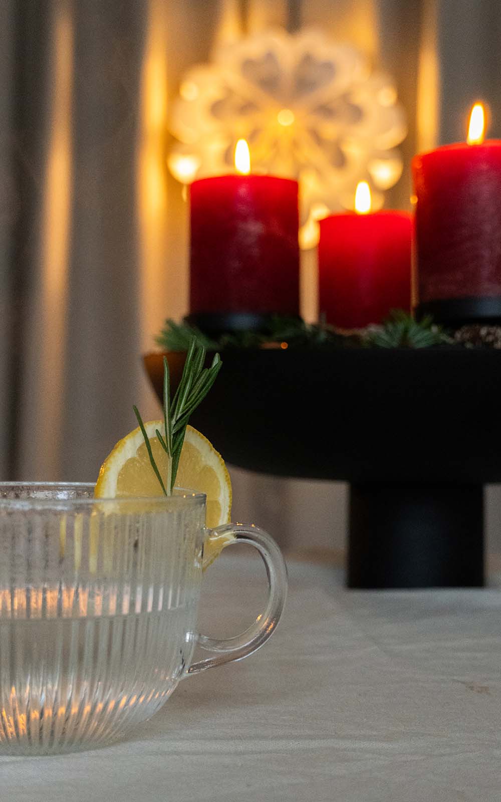 Glas mit Rosmarin-Zitronen-Tee, garniert mit einer Zitronenscheibe und Rosmarinzweig, im Hintergrund leuchtende rote Kerzen auf einem Adventskranz. Glas mit Rosmarin-Zitronen-Tee, garniert mit einer Zitronenscheibe und Rosmarinzweig, im Hintergrund leuchtende rote Kerzen auf einem Adventskranz.