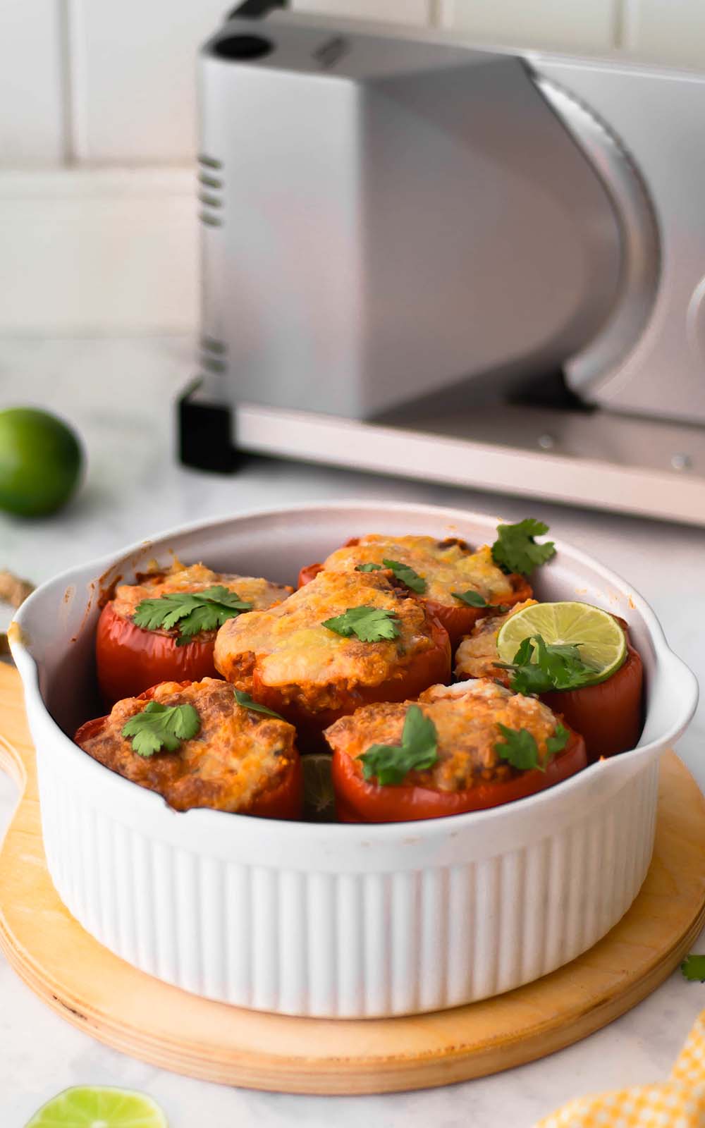 Auflaufform mit mehreren gefüllten Ofenpaprika, überbacken mit Käse und garniert mit frischem Koriander und Limettenscheiben. Im Hintergrund ein elektrischer Allesschneider Auflaufform mit mehreren gefüllten Ofenpaprika, überbacken mit Käse und garniert mit frischem Koriander und Limettenscheiben. Im Hintergrund ein elektrischer Allesschneider