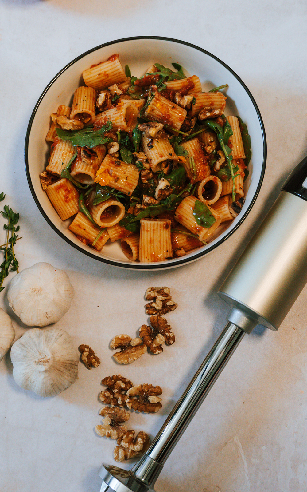 Ein Teller mit Rigatoni-Nudeln in Tomatensauce, angerichtet mit frischem Rucola, Walnüssen und Kräutern. Daneben liegen frische Knoblauchknollen, lose Walnusshälften und ein silberfarbener Stabmixer auf einer hellen Küchenoberfläche