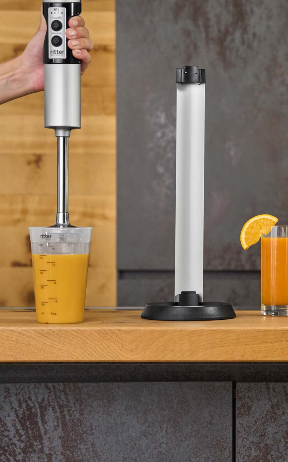 A picture of a silver hand blender mixing a juice in a measuring cup and a glass of squeezed orange juice standing in a kitchen. A picture of a silver hand blender mixing a juice in a measuring cup and a glass of squeezed orange juice standing in a kitchen.