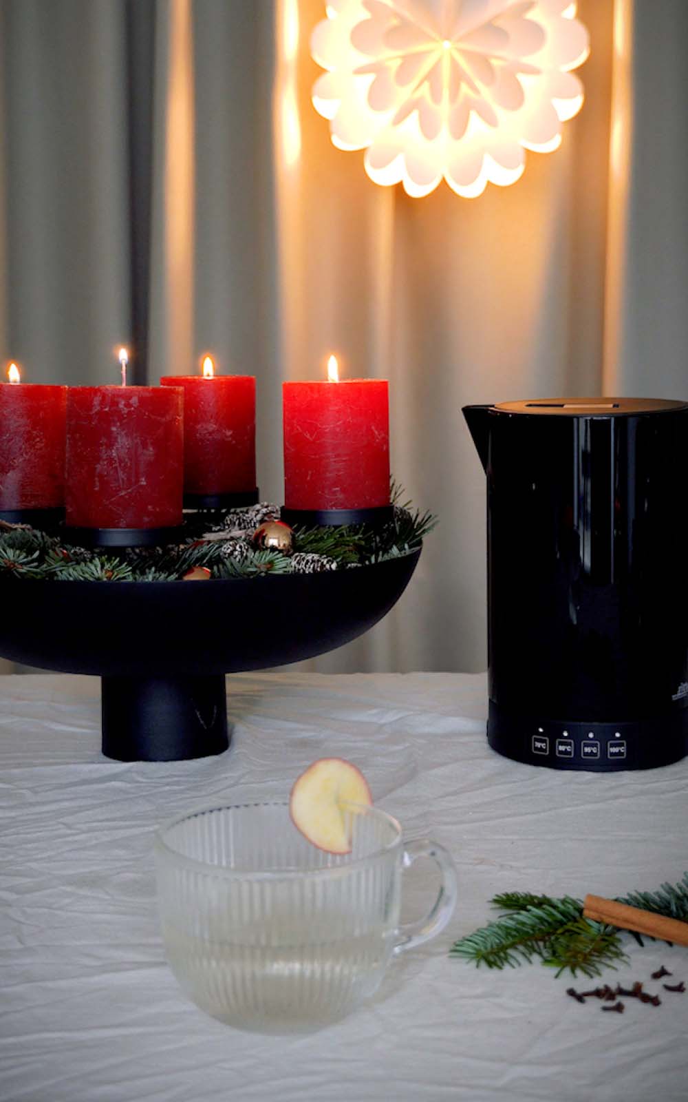Glasbecher mit heißer Apfel-Zimt-Infusion, garniert mit einer Apfelscheibe, daneben Tannenzweige und Zimtstange; im Hintergrund Adventskranz mit roten Kerzen und ein schwarzer Wasserkocher. Glasbecher mit heißer Apfel-Zimt-Infusion, garniert mit einer Apfelscheibe, daneben Tannenzweige und Zimtstange; im Hintergrund Adventskranz mit roten Kerzen und ein schwarzer Wasserkocher.