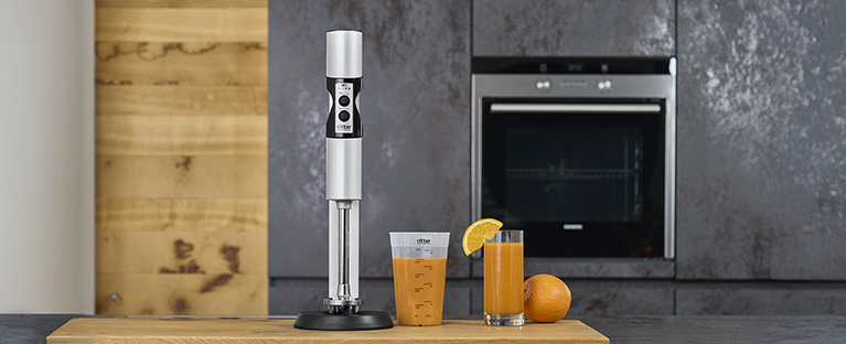 A picture of a silver hand blender, a filled measuring jug and a glass filled with juice in a kitchen.