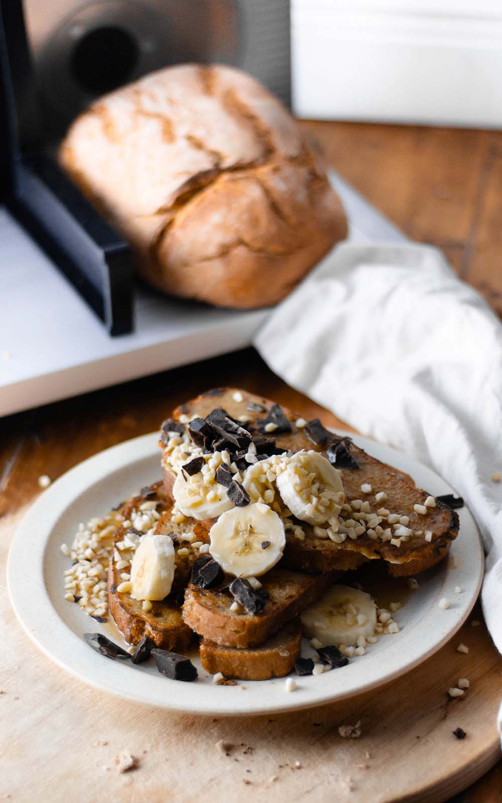 Goldbraun gebratener French Toast auf einem Teller, belegt mit frischen Bananenscheiben, gehackten Nüssen und dunklen Schokoladenstückchen, im Hintergrund ein Laib Brot. Goldbraun gebratener French Toast auf einem Teller, belegt mit frischen Bananenscheiben, gehackten Nüssen und dunklen Schokoladenstückchen, im Hintergrund ein Laib Brot.