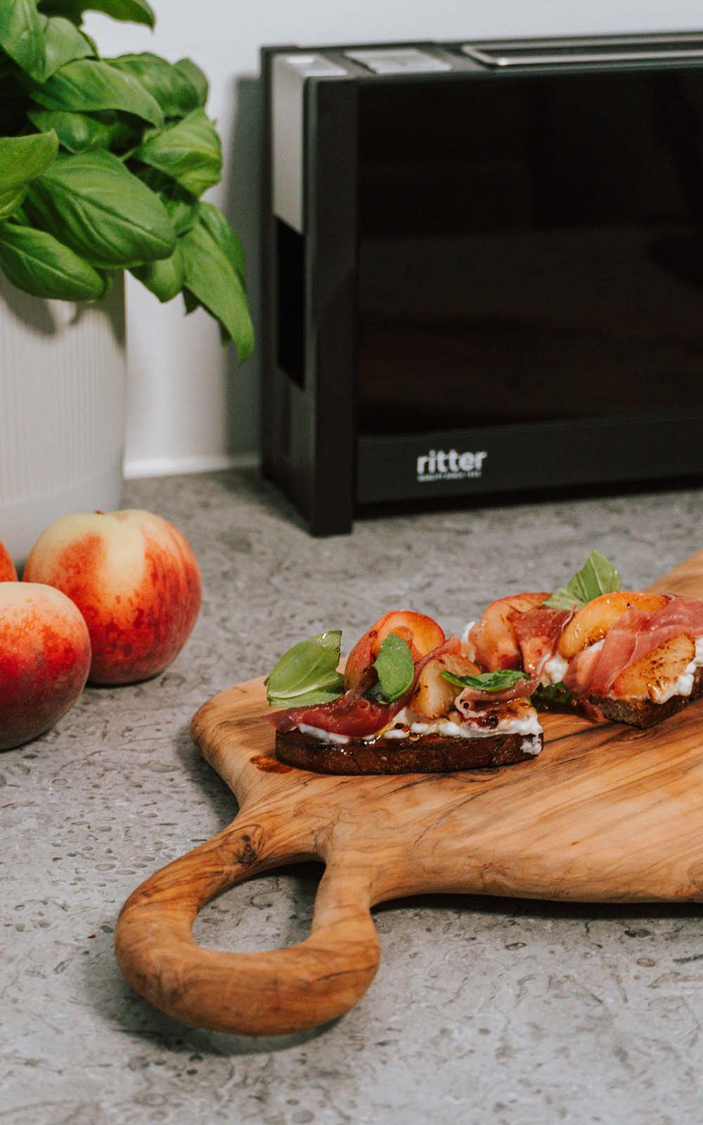 Zwei Sommer-Toasts mit frischen Pfirsichen, Schinken, Frischkäse und Basilikum auf einem rustikalen Holzbrett. Daneben stehen Pfirsiche und ein Topf mit Basilikum, im Hintergrund eine Küchenmaschine