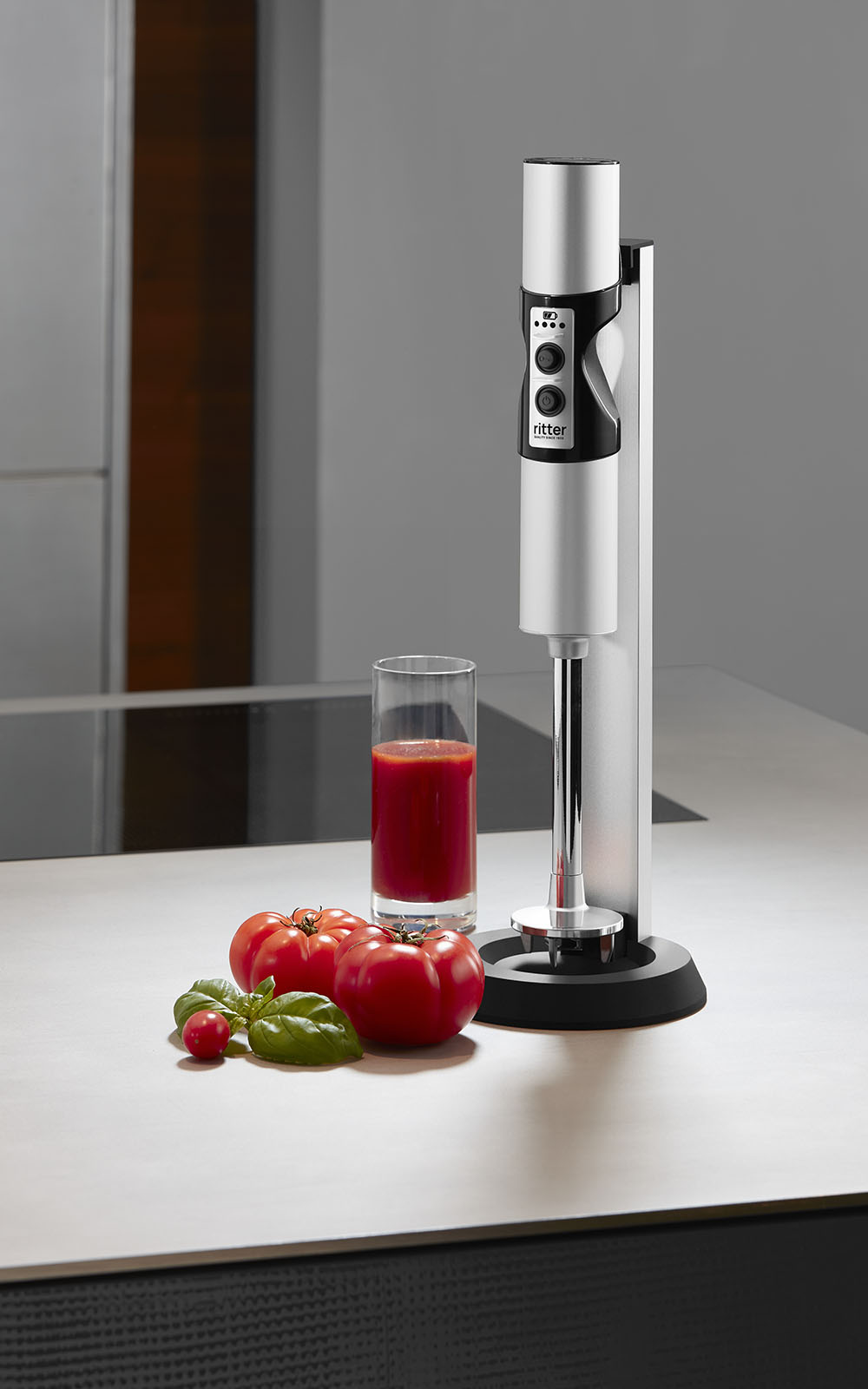 A picture of a silver hand blender standing next to a glass of tomato juice in a kitchen. A picture of a silver hand blender standing next to a glass of tomato juice in a kitchen.