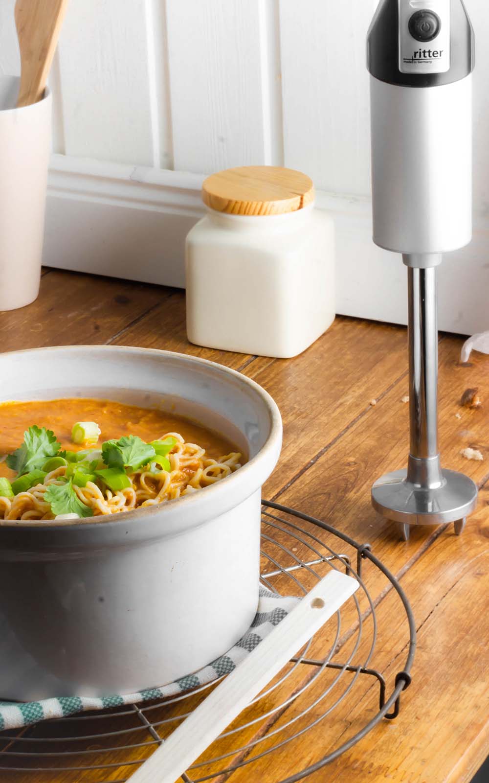 Schüssel mit cremiger Thai-Suppe, serviert mit Nudeln, Frühlingszwiebeln und frischem Koriander, auf einem Holztisch mit Stabmixer und Küchenutensilien im Hintergrund. Schüssel mit cremiger Thai-Suppe, serviert mit Nudeln, Frühlingszwiebeln und frischem Koriander, auf einem Holztisch mit Stabmixer und Küchenutensilien im Hintergrund.