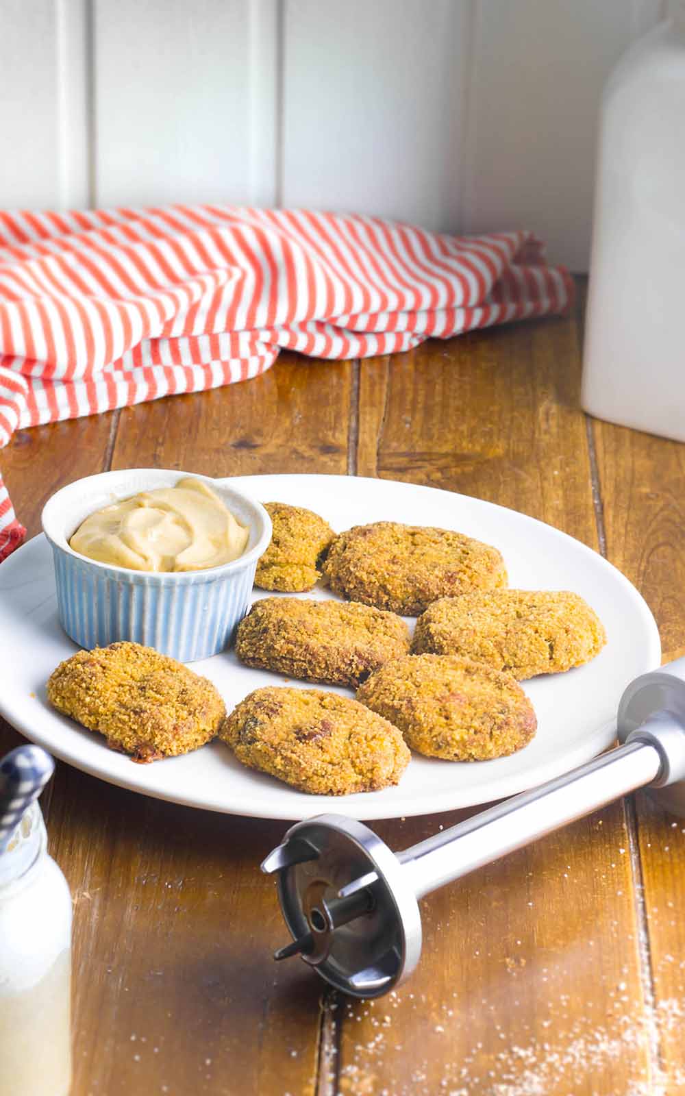 Ein Teller mit knusprigen, goldbraun gebackenen Veggie-Nuggets, serviert mit einer Schale cremigem Dip. Daneben liegt ein Stabmixer, im Hintergrund ein rot-weiß gestreiftes Tuch auf einem Holztisch