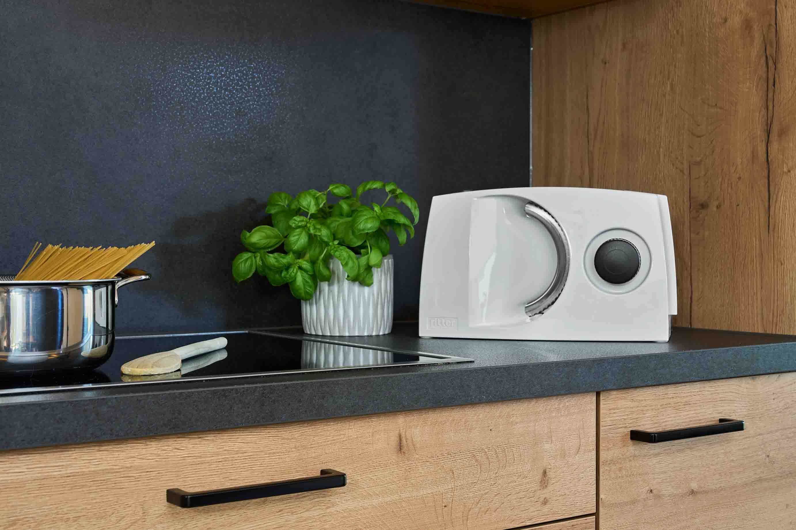 White food slicer on a dark kitchen worktop with a pot of basil and spaghetti in the pot.