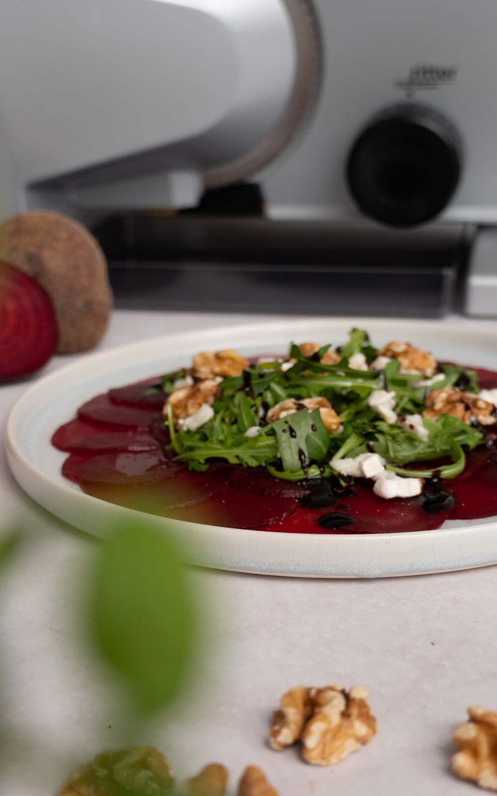 Carpaccio aus Rote-Bete-Scheiben, garniert mit Rucola, Walnüssen, Ziegenkäse und Balsamico-Creme auf einem weißen Teller, angerichtet vor einem ritter Allesschneider