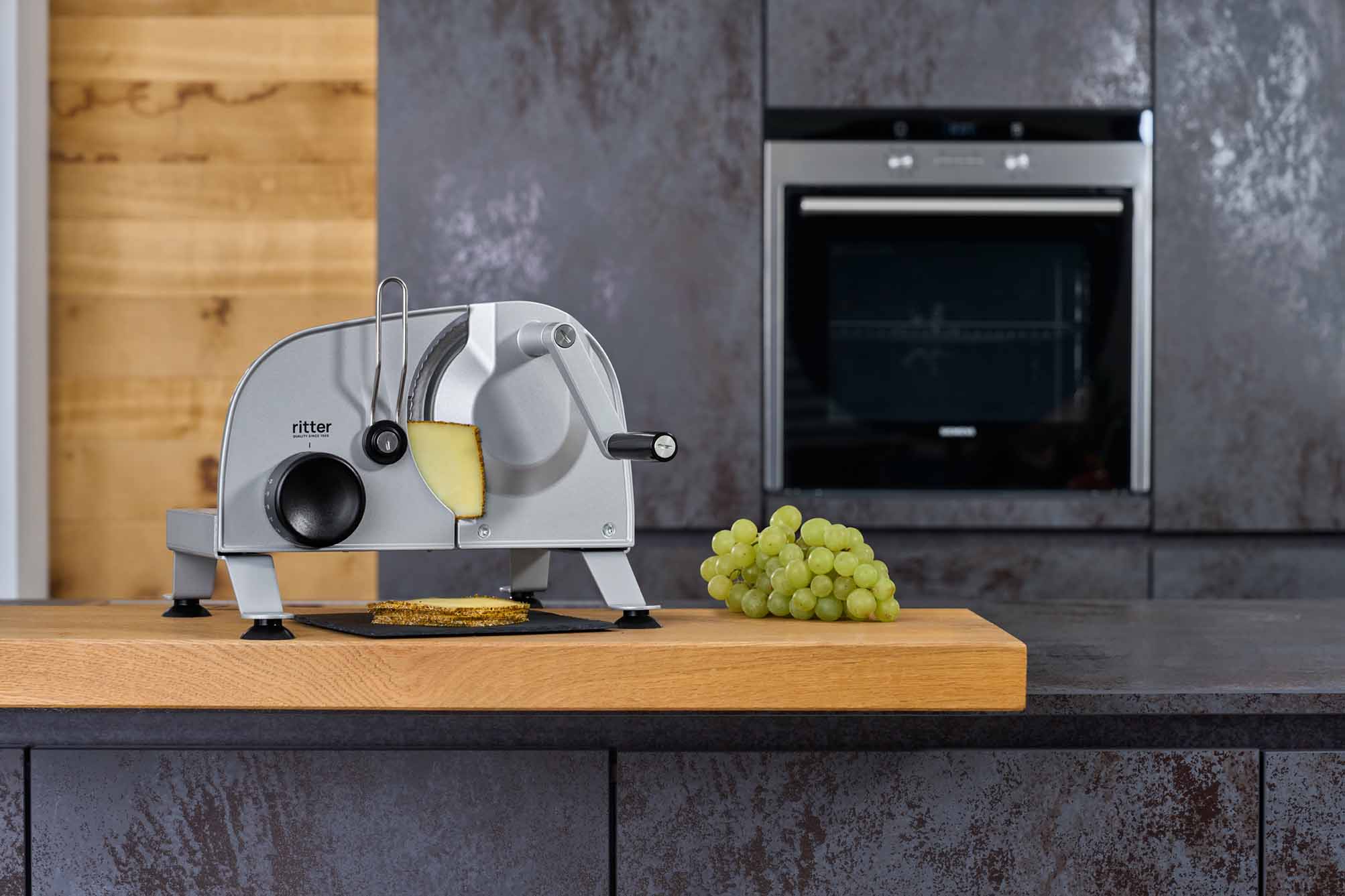 Silberner manueller Allesschneider auf Holzarbeitsplatte mit geschnittenem Käse und grünem Traubenbündel in moderner Küche