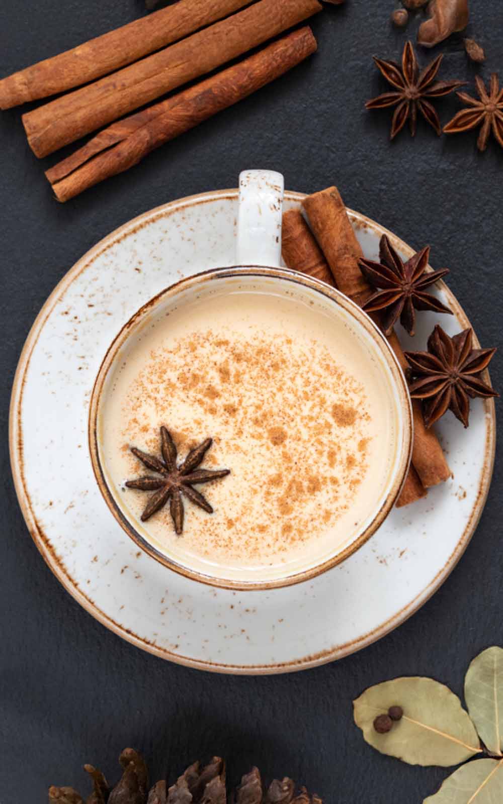 Tasse cremiger Chai Latte mit Zimt bestäubt und Sternanis als Dekoration, serviert auf Untertasse; daneben Zimtstangen, Sternanis, Lorbeerblätter und Gewürze auf dunkler Oberfläche. Tasse cremiger Chai Latte mit Zimt bestäubt und Sternanis als Dekoration, serviert auf Untertasse; daneben Zimtstangen, Sternanis, Lorbeerblätter und Gewürze auf dunkler Oberfläche.