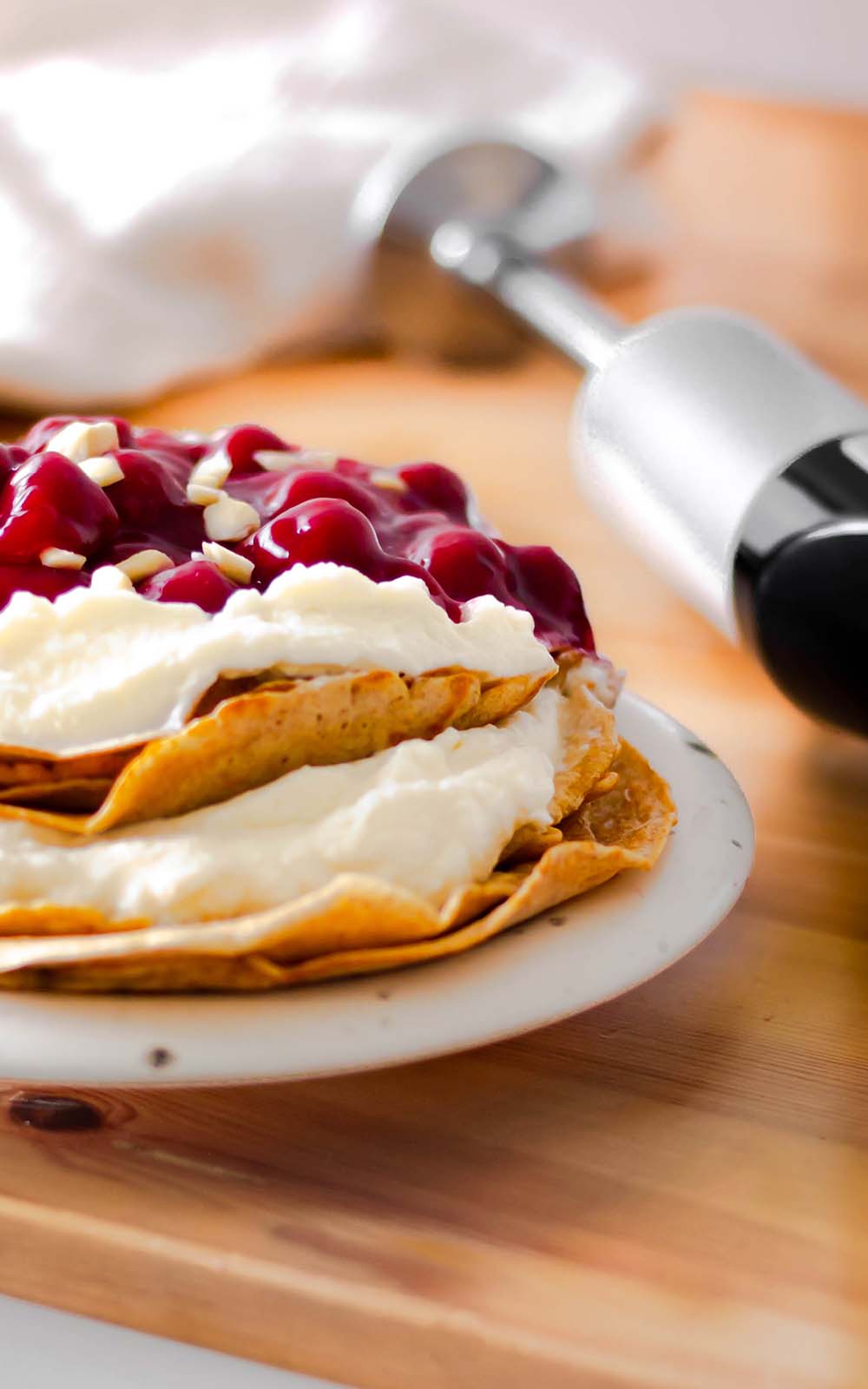 Gestapelte Pfannkuchen, gefüllt mit cremiger Sahne und getoppt mit einer fruchtigen Kirschsoße und gehackten Mandeln, serviert auf einem weißen Teller. Im Hintergrund liegt ein Schneebesen auf einem Holzbrett. Gestapelte Pfannkuchen, gefüllt mit cremiger Sahne und getoppt mit einer fruchtigen Kirschsoße und gehackten Mandeln, serviert auf einem weißen Teller. Im Hintergrund liegt ein Schneebesen auf einem Holzbrett.