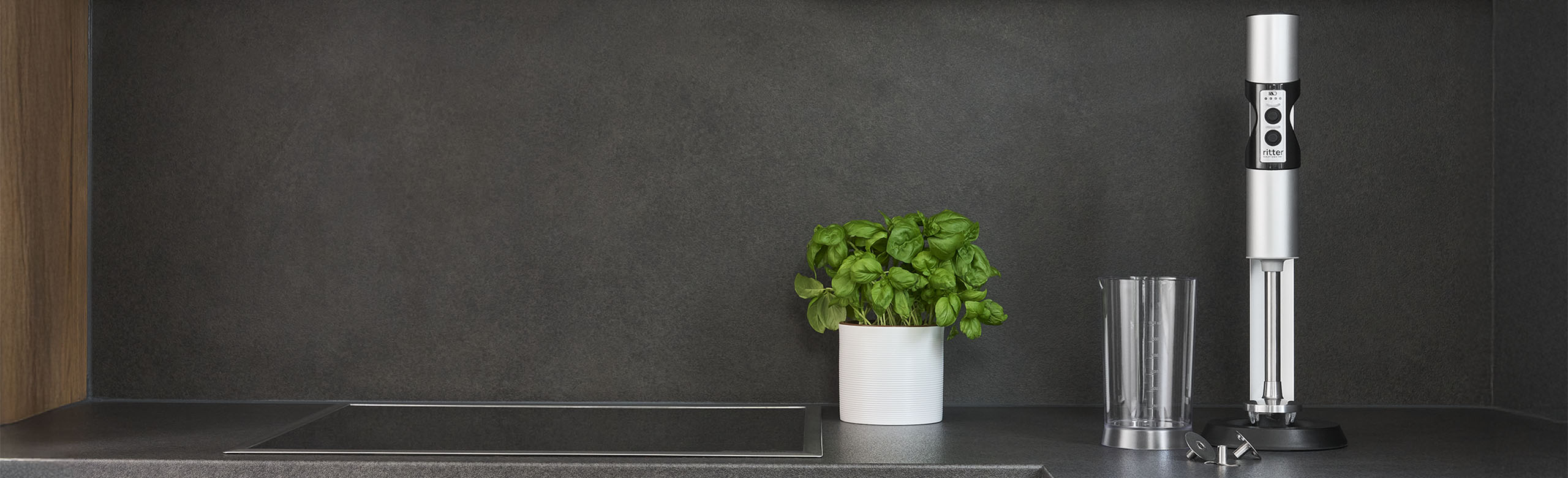 Ein Bild von einem silbernen Stabmixer, einem gefüllten Messbecher und einem, mit Saft gefüllten, Glas, in einer Küche.