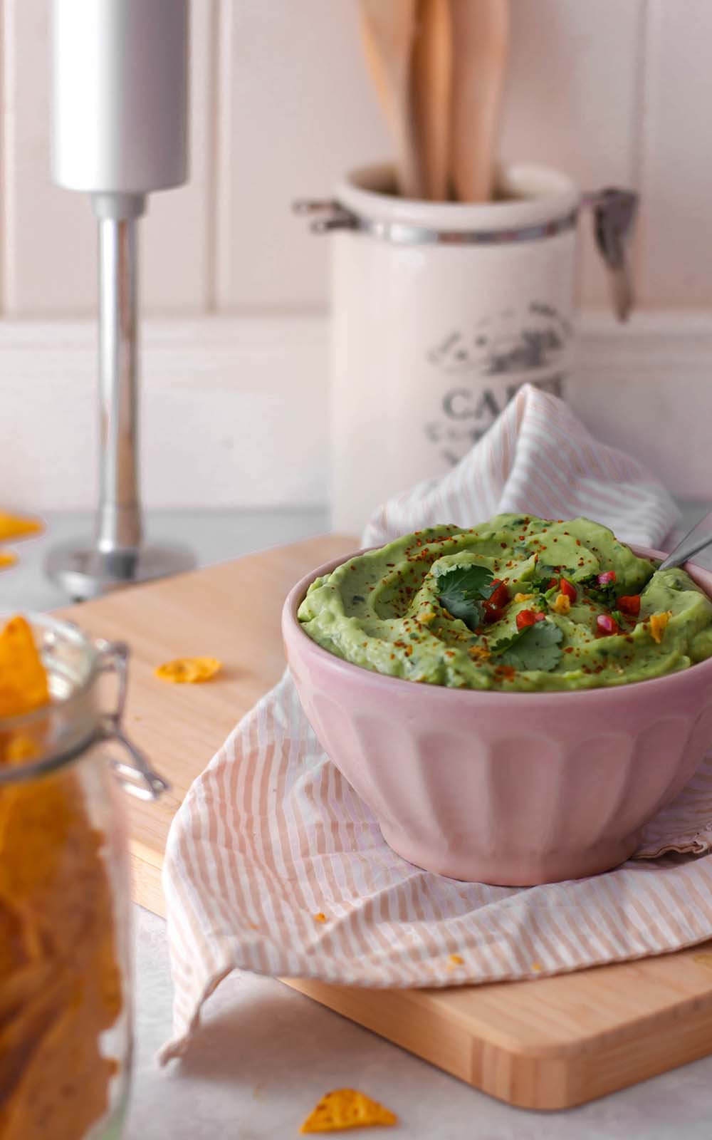 Eine rosa Schüssel gefüllt mit cremigem Avocado-Dip, garniert mit frischen Kräutern und gehacktem Chili. Im Vordergrund sind Tortilla-Chips im Glas zu sehen, die sich perfekt zum Dippen eignen. Der Dip steht auf einem Holzbrett mit gestreiftem Tuch Eine rosa Schüssel gefüllt mit cremigem Avocado-Dip, garniert mit frischen Kräutern und gehacktem Chili. Im Vordergrund sind Tortilla-Chips im Glas zu sehen, die sich perfekt zum Dippen eignen. Der Dip steht auf einem Holzbrett mit gestreiftem Tuch