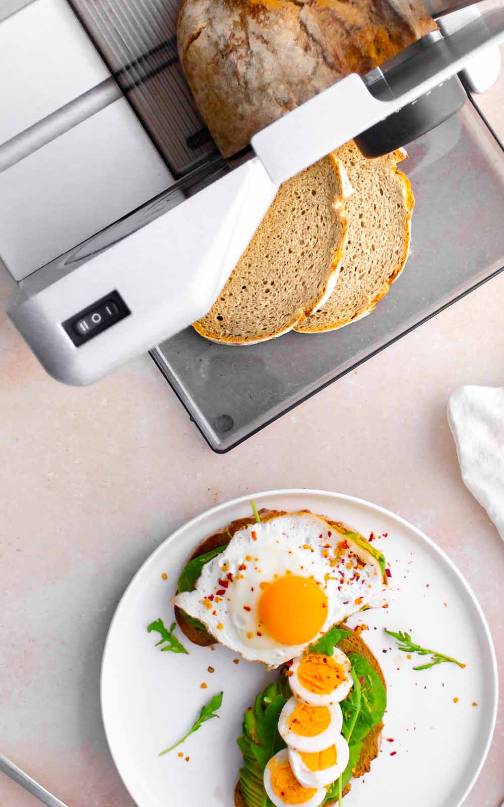 Frisches Brot wird mit einer Brotschneidemaschine in Scheiben geschnitten. Auf einem Teller liegt ein belegtes Avocado-Brot mit Spinat, gekochten Eierscheiben und einem Spiegelei, garniert mit Rucola und Chiliflocken. Frisches Brot wird mit einer Brotschneidemaschine in Scheiben geschnitten. Auf einem Teller liegt ein belegtes Avocado-Brot mit Spinat, gekochten Eierscheiben und einem Spiegelei, garniert mit Rucola und Chiliflocken.
