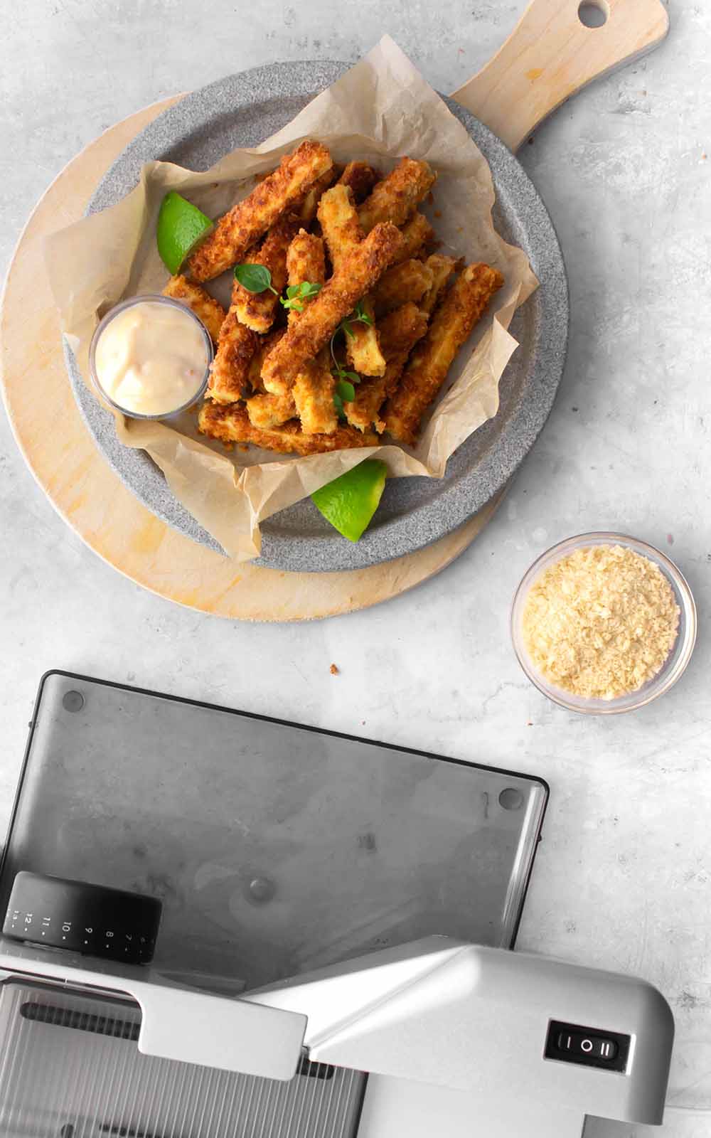 Knusprig gebackene Tofu-Sticks auf Backpapier, serviert auf einem Teller mit Dip und Limettenstück. Daneben eine kleine Schale mit Panko-Bröseln und im Vordergrund eine silberne Küchenmaschine