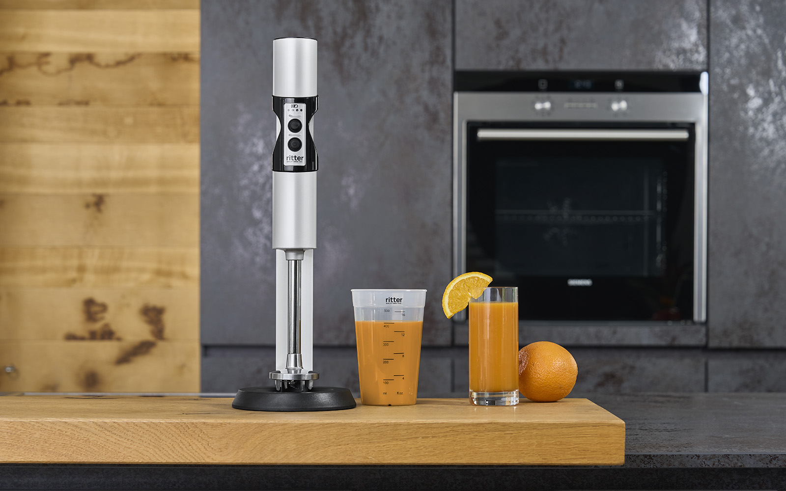 Ein Bild von einem silbernen Stabmixer, einem gefüllten Messbecher und einem, mit Saft gefüllten, Glas, in einer Küche.