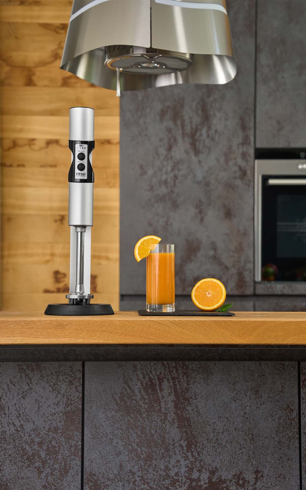 Ein Bild eines silbernen Stabmixers der neben einem Glas Orangensaft, in einer Küche steht. Ein Bild eines silbernen Stabmixers der neben einem Glas Orangensaft, in einer Küche steht.