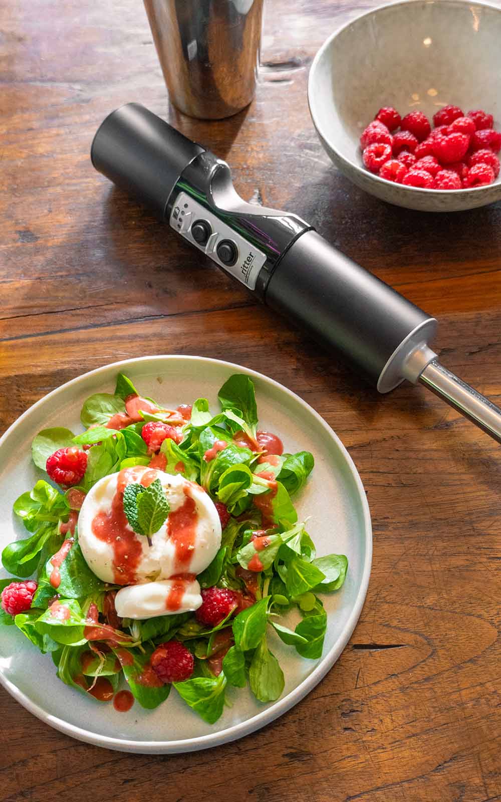 Ein Teller mit frischem Feldsalat, garniert mit Burrata, frischen Himbeeren und einem leuchtend roten Himbeerdressing, verziert mit einem Minzblatt. Daneben steht eine Schale mit Himbeeren und ein Pürierstab Ein Teller mit frischem Feldsalat, garniert mit Burrata, frischen Himbeeren und einem leuchtend roten Himbeerdressing, verziert mit einem Minzblatt. Daneben steht eine Schale mit Himbeeren und ein Pürierstab