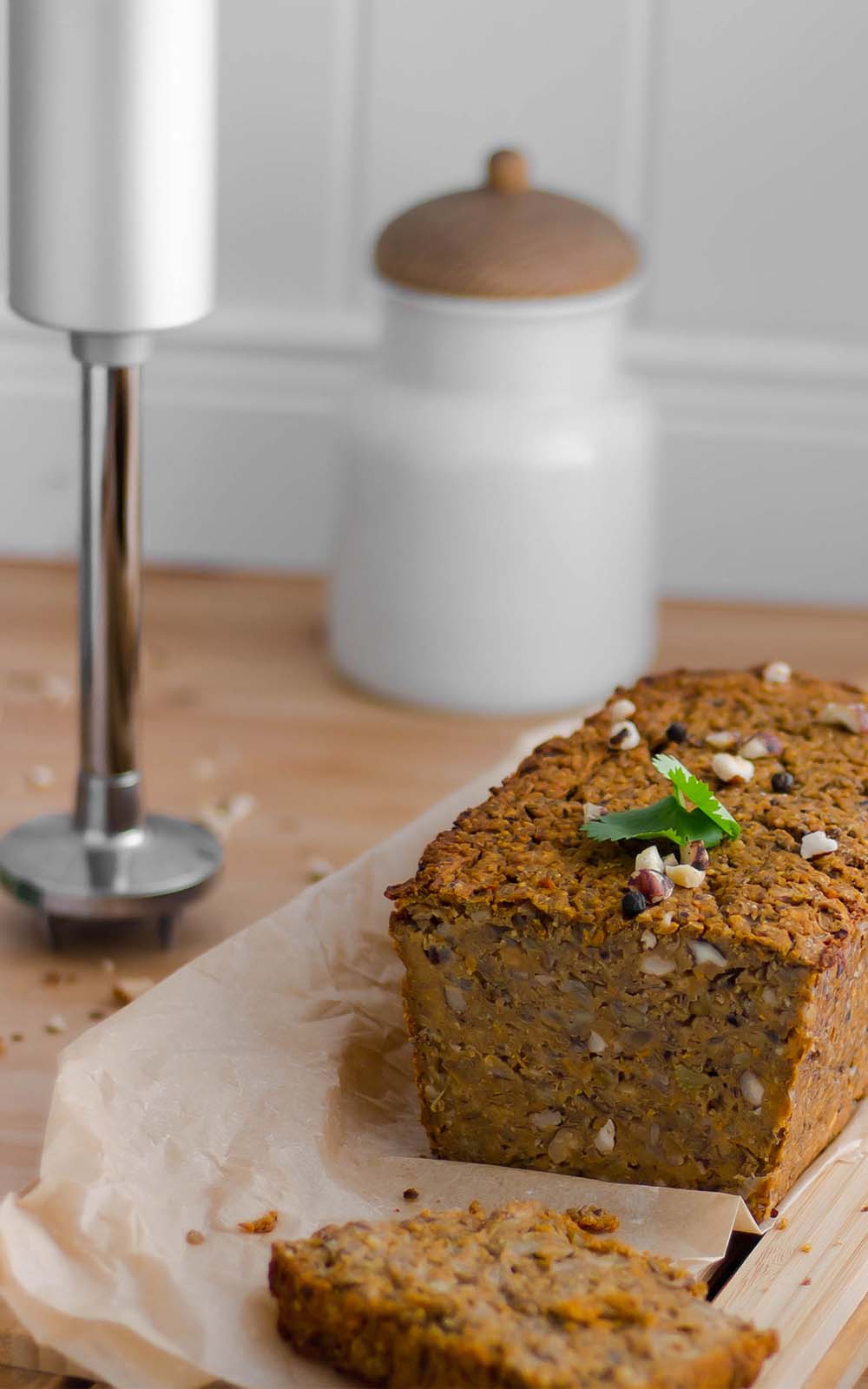 Vegetarischer Nussbraten auf Backpapier, mit gehackten Nüssen und frischen Kräutern garniert. Daneben liegt eine abgeschnittene Scheibe, im Hintergrund ein Stabmixer und ein weißes Vorratsgefäß