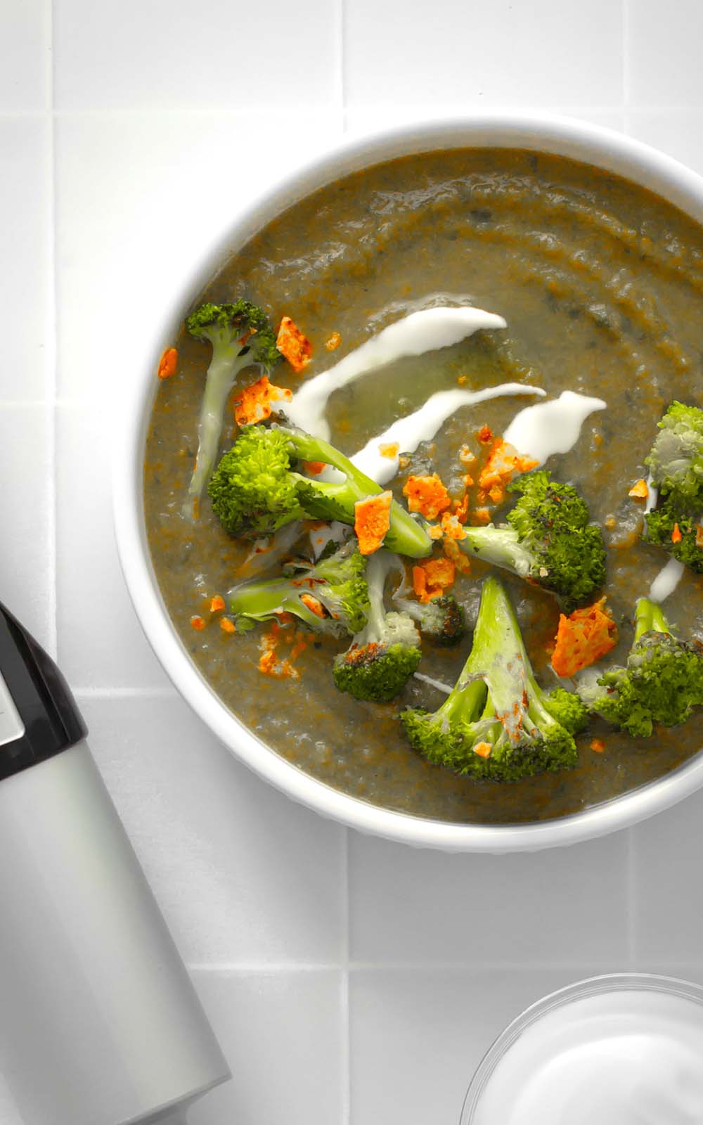 Grüne cremige Suppe in einer Schüssel, garniert mit Brokkoli-Röschen, orangenfarbigen Toppings und einem Klecks weißer Creme, daneben ein Stabmixer und ein Glas. Grüne cremige Suppe in einer Schüssel, garniert mit Brokkoli-Röschen, orangenfarbigen Toppings und einem Klecks weißer Creme, daneben ein Stabmixer und ein Glas.