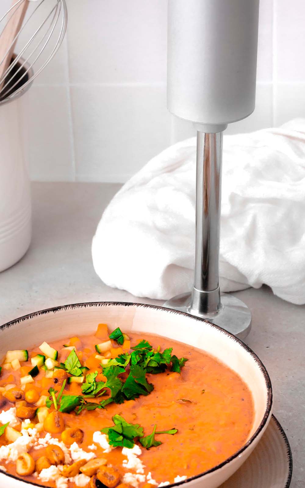 Gazpacho in einer Schüssel, garniert mit gehacktem Gemüse, frischen Kräutern, gerösteten Nüssen und etwas Käse, daneben ein Stabmixer auf einer Küchenarbeitsfläche. Gazpacho in einer Schüssel, garniert mit gehacktem Gemüse, frischen Kräutern, gerösteten Nüssen und etwas Käse, daneben ein Stabmixer auf einer Küchenarbeitsfläche.