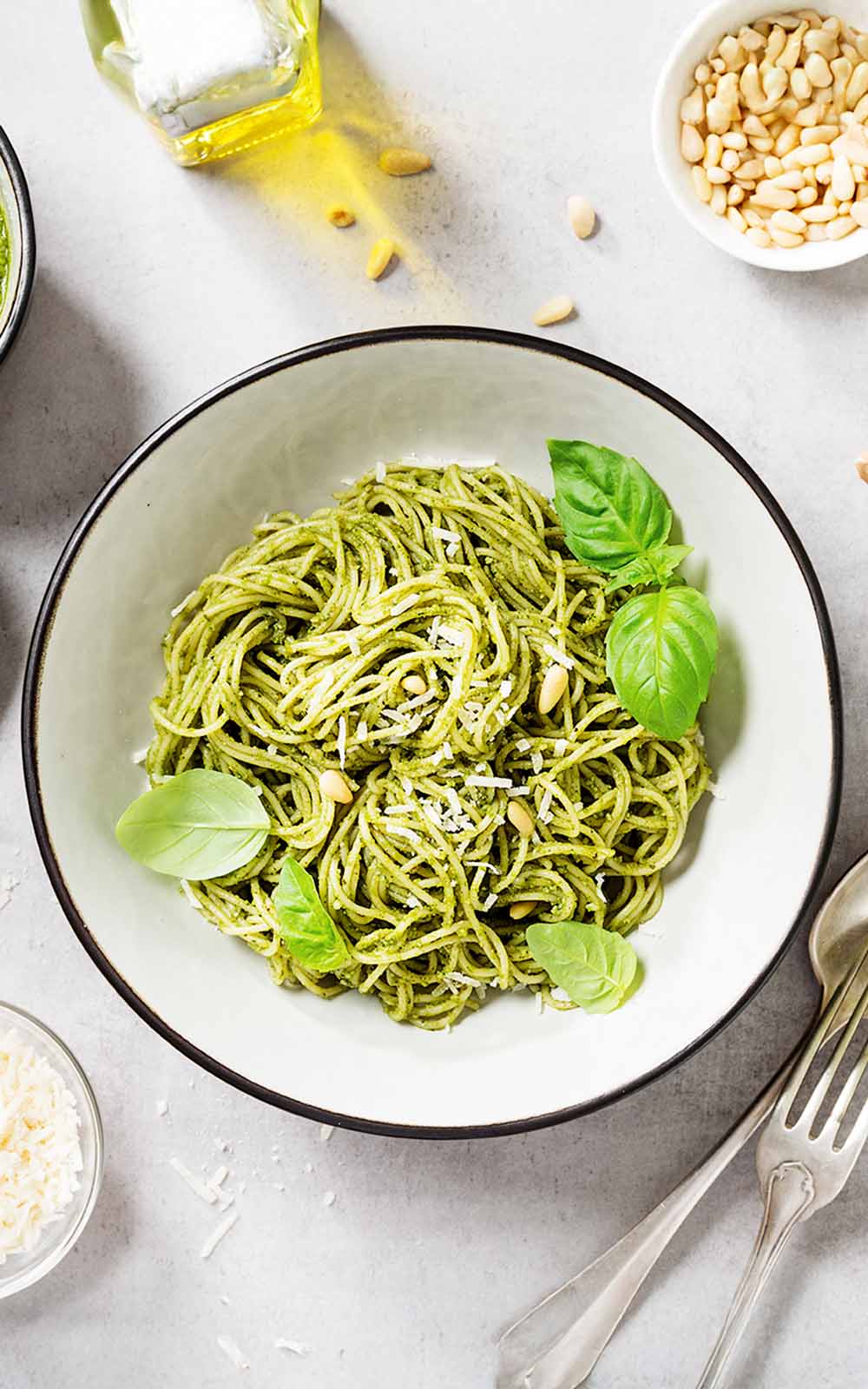 Schüssel mit Spaghetti in grünem Pesto, garniert mit frischem Basilikum, Pinienkernen und geriebenem Parmesan. Daneben liegen Besteck, Olivenöl und kleine Schalen mit Zutaten