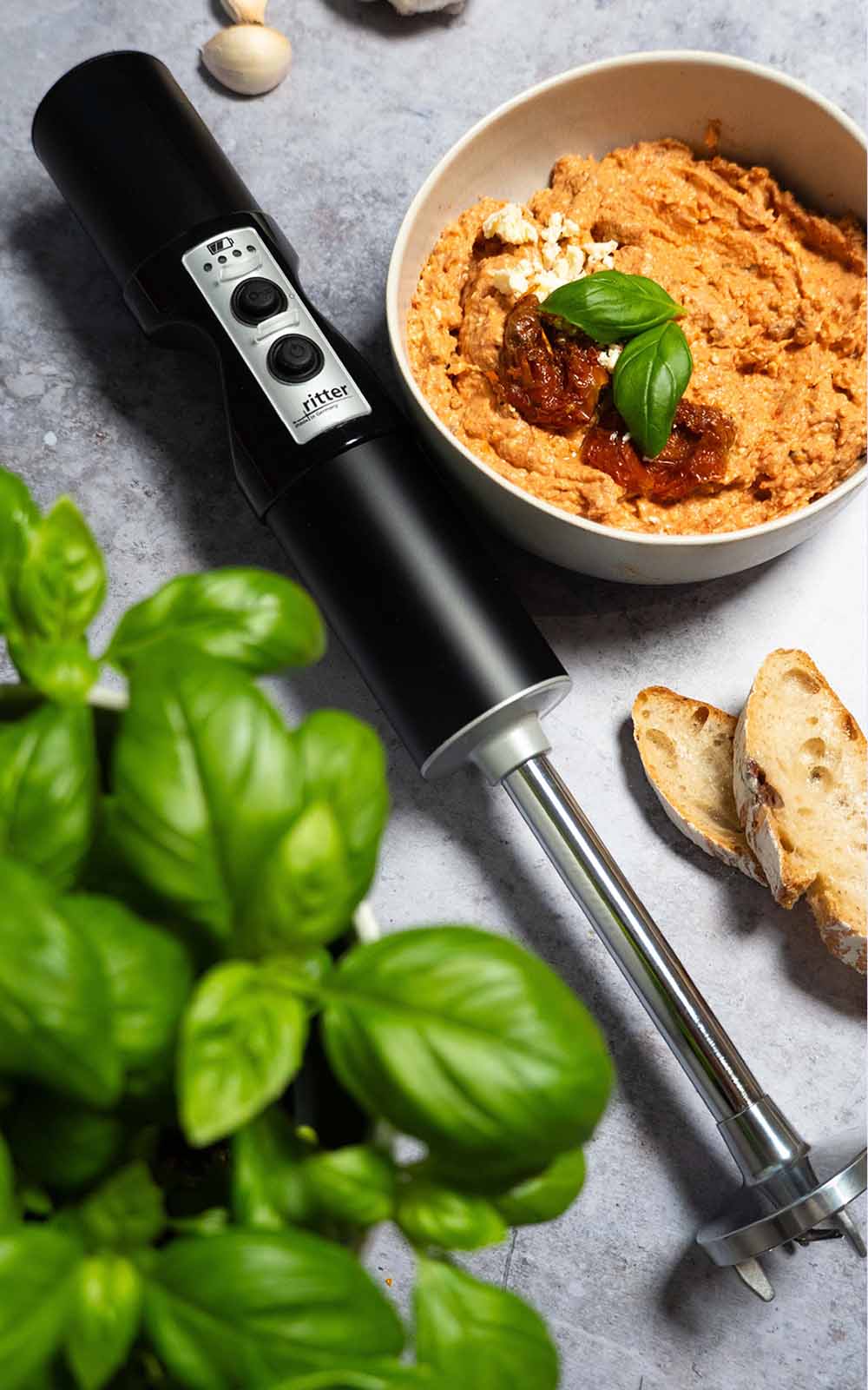 Eine Schüssel mit cremigem Tomaten-Feta-Dip, garniert mit getrockneten Tomaten, zerbröckeltem Feta und frischem Basilikum. Daneben liegen knusprige Brotscheiben und ein Pürierstab, im Vordergrund frische Basilikumblätter. Eine Schüssel mit cremigem Tomaten-Feta-Dip, garniert mit getrockneten Tomaten, zerbröckeltem Feta und frischem Basilikum. Daneben liegen knusprige Brotscheiben und ein Pürierstab, im Vordergrund frische Basilikumblätter.