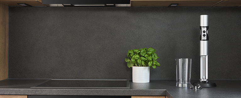 Ein Bild von einem silbernen Stabmixer, einem gefüllten Messbecher und einem, mit Saft gefüllten, Glas, in einer Küche.