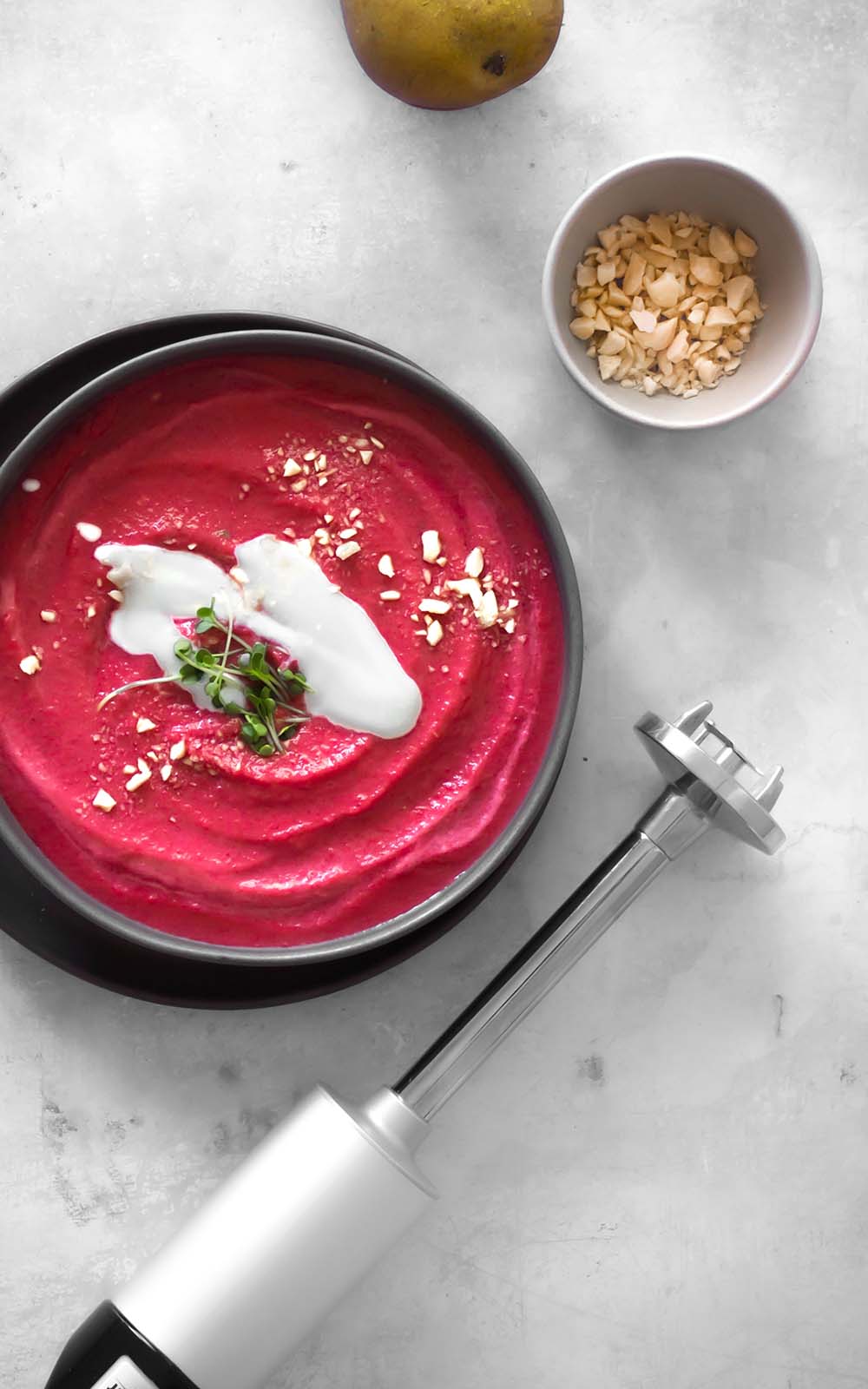 Schwarze Schüssel mit cremiger Rote-Bete-Suppe, garniert mit Kokosjoghurt, frischen Sprossen und gehackten Nüssen, daneben ein Stabmixer auf hellem Untergrund. Schwarze Schüssel mit cremiger Rote-Bete-Suppe, garniert mit Kokosjoghurt, frischen Sprossen und gehackten Nüssen, daneben ein Stabmixer auf hellem Untergrund.