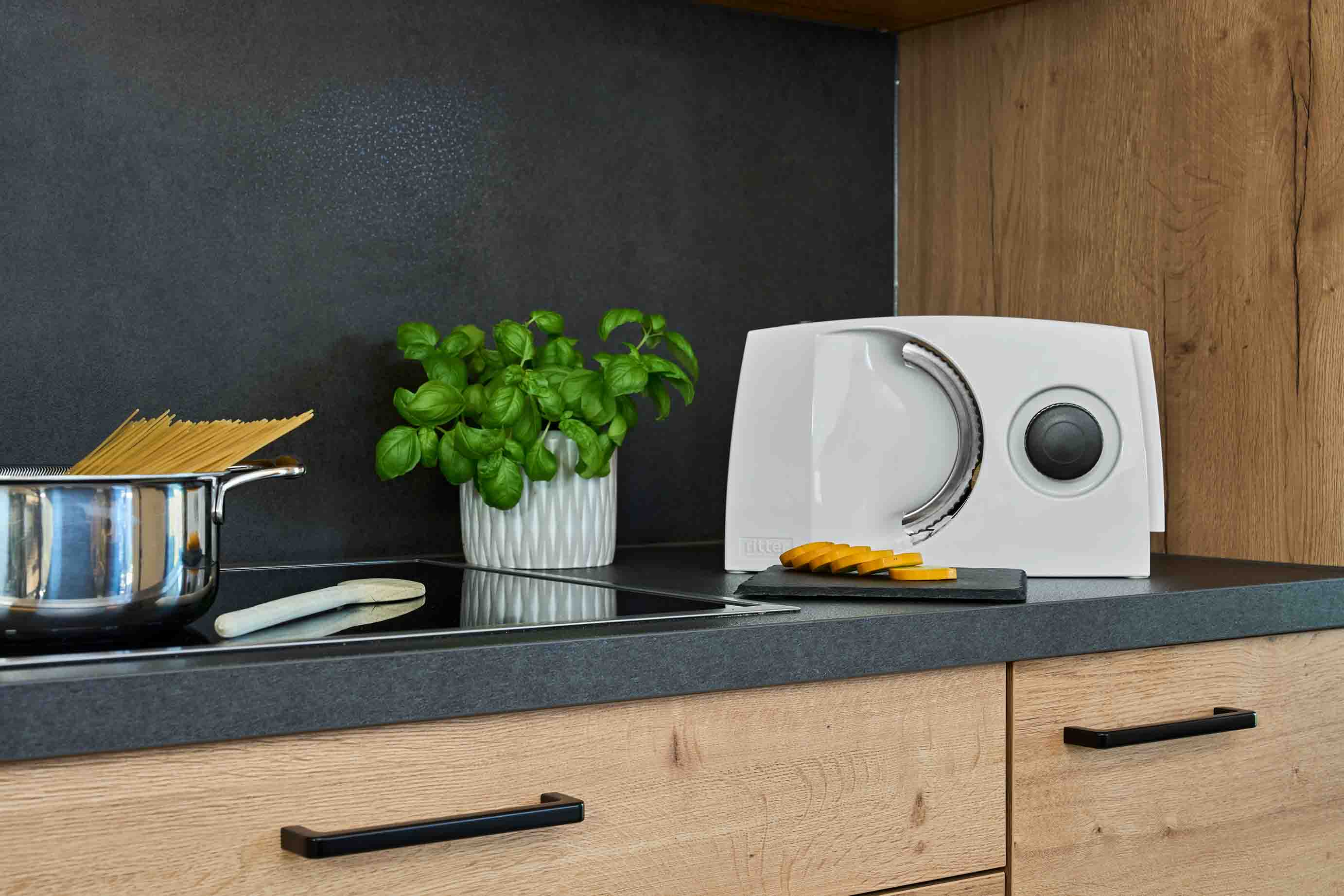 White food slicer on a dark kitchen worktop with sliced food, a pot of basil and spaghetti in a pot.