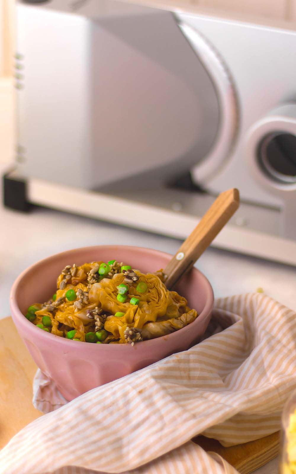 Schüssel mit gebackenem Ofenspitzkohl, garniert mit grünen Frühlingszwiebeln und Samen. Im Hintergrund steht ein elektrischer Allesschneider