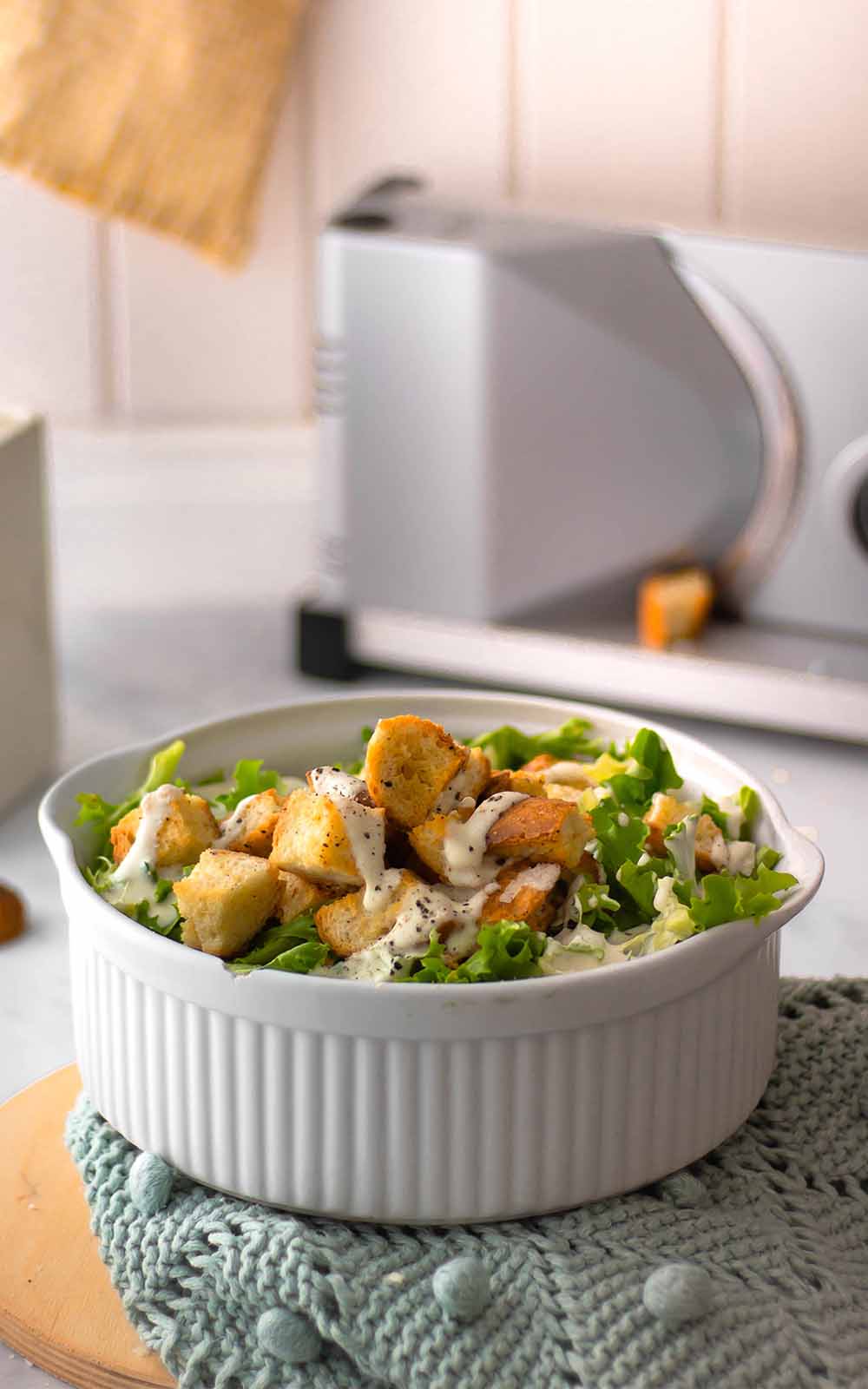 Weiße Schüssel mit frischem Brotsalat aus grünem Blattsalat, knusprigen Croutons und cremigem Dressing, serviert auf einem gestrickten Untersetzer. Weiße Schüssel mit frischem Brotsalat aus grünem Blattsalat, knusprigen Croutons und cremigem Dressing, serviert auf einem gestrickten Untersetzer.